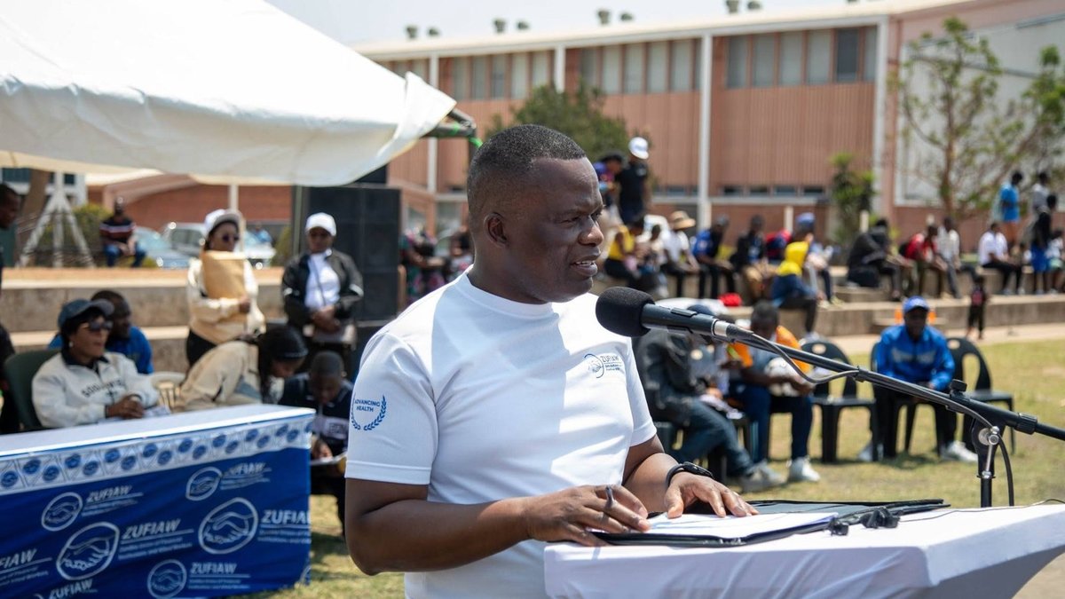 Labour Commissioner Mr. Givens Muntengwa speaking at an outdoor podium in a white ZUFIAW shirt, with festival participants seated behind him.